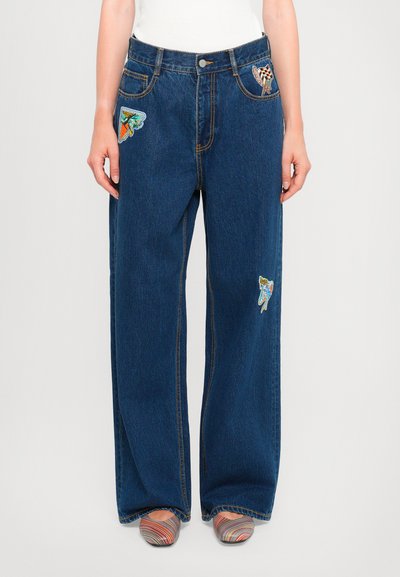 Jeans en denim bleu à jambes larges avec des patchs colorés brodés sur les poches avant et des coutures contrastantes. Portés avec des chaussures multicolores.