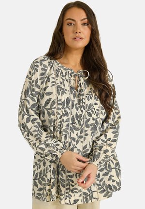 Kobieta o długich brązowych włosach, ubrana w kremową bluzkę z czarnym liściastym wzorem i wiązaniami przy dekolcie, stoi na jednolitym tle.