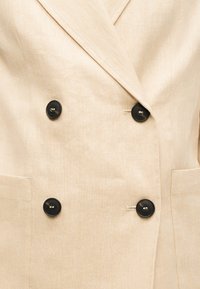 Μπεζ διπλόπατο σακάκι με μαύρα κουμπιά και δύο μπροστινές τσέπες, κατασκευασμένο από υφή υφάσματος.