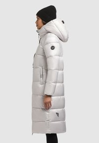 Hosszú, bélelt ezüstszínű kabát kapucnival, elülső cipzárral, két oldalzsebbel és egy logóval a vállán. Fekete sapka viselve.