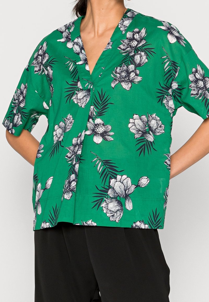 Blouse verte à manches courtes avec imprimé floral noir et blanc portée par une personne les mains sur les hanches, associée à un pantalon noir.