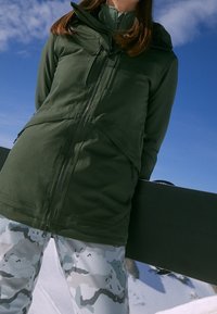 Giacca leggera verde con chiusura a zip e cappuccio; presenta tasche laterali e uno strato interno testurizzato. Abbinata a pantaloni grigi camo con motivo.