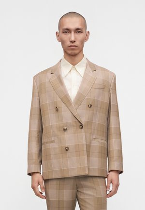 Hombre con cabeza rapada vistiendo un traje cruzado de cuadros beige y marrón claro y una camisa crema abotonada, de pie frente a un fondo liso.