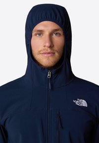 Marinblå huvjacka med elastisk huva, dragkedja fram och broderad logotyp. Har en dragkedjeficka på vänster bröst.