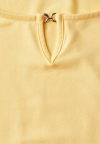 Žlutý textilní top s designem do V a malým kovovým kroužkem na výstřihu, vyrobený z hladkého a elastického materiálu.