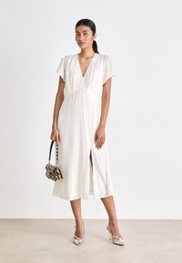 Biała, średniej długości sukienka z krótkimi, falbaniaste rękawami, głębokim dekoltem w literę V i bocznym rozcięciem. Noszona z torebką w wzory i metalicznymi szpilkami.