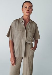 Camicia a maniche corte di colore taupe, con bottoni e due tasche frontali; presenta una vestibilità rilassata e un tessuto leggero e testurizzato. Abbinata a pantaloni coordinati.