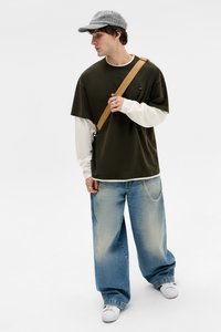 Olivgrünes, oversized T-Shirt über einem weißen Langarmshirt, kombiniert mit weit geschnittenen hellblauen Jeans mit Kettendetails und einer grauen Kappe.