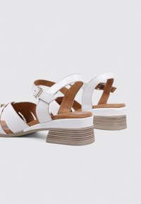 Sandalias blancas con diseño de tiras cruzadas, plantillas de cuero marrón y un tacón apilado y texturizado. Presentan hebillas ajustables y un acabado suave.