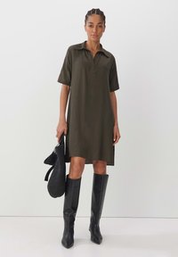 Olivenfarbendes Kragenkleid mit kurzen Ärmeln, lässiger Schnitt, seitliche Schlitze; kombiniert mit knielangen schwarzen Stiefeln und einer strukturierten schwarzen Handtasche.