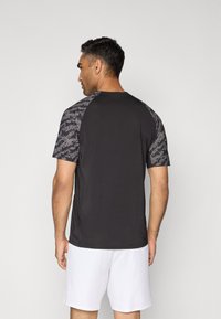 Sort atletisk T-shirt med korte ærmer, grå mønstrede raglan-skuldrer og glat tekstur, parret med hvide shorts.