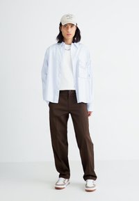 Chemise bleu clair à boutons, t-shirt blanc, pantalon marron, baskets crème avec accents marron, et casquette beige avec texte. Tenue décontractée, design minimaliste.