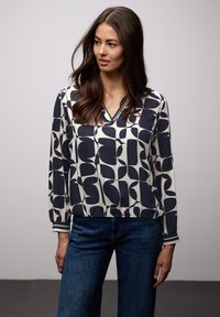 Femme aux longs cheveux bruns portant un chemisier à motifs abstraits noir et blanc et un jean bleu, debout devant un fond clair uni.