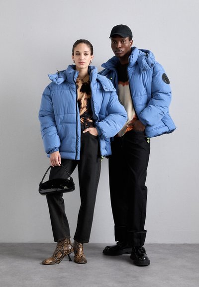 Hellblaue Steppjacken mit Kapuzen, präsentiert von zwei Models. Einer trägt einen gemusterten Pullover und dunkle Hosen; der andere trägt eine Mütze und schwarze Schuhe.