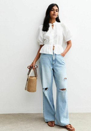 White puff-sleeve blouse with a tie-front closure, paired with light blue wide-leg ripped jeans. The model holds a circular woven bag.