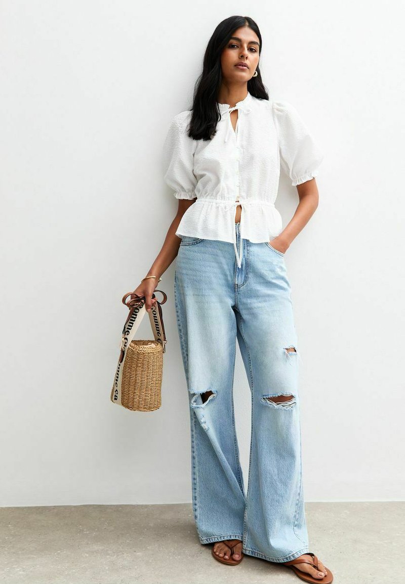 Vit blus med puffärmar och knytning framtill, i kombination med ljusblå vida rivna jeans. Modellen håller en rund vävd väska.