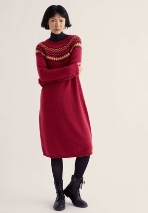 Femme aux cheveux courts noirs portant une robe en maille rouge à motifs, des leggings noirs et des bottes noires à lacets, debout les bras croisés.