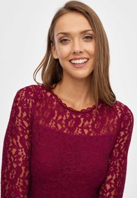 Top de encaje burdeos con patrones florales, escote con forma de ondas y mangas largas, con una capa inferior suave.