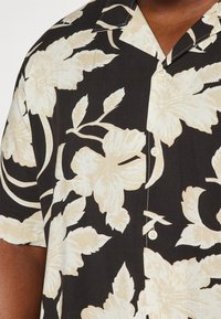 Camisa negra de manga corta con grandes patrones florales beiges, cuello con muesca y botones blancos a lo largo de la tapeta. Textura de tela suave.