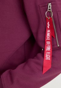 Sweat-shirt bordeaux avec une poche zippée comportant une étiquette en ruban rouge portant la mention "ENLEVER AVANT LE VOL" en blanc. Tissu texturé. Détail de fermeture éclair métallique.