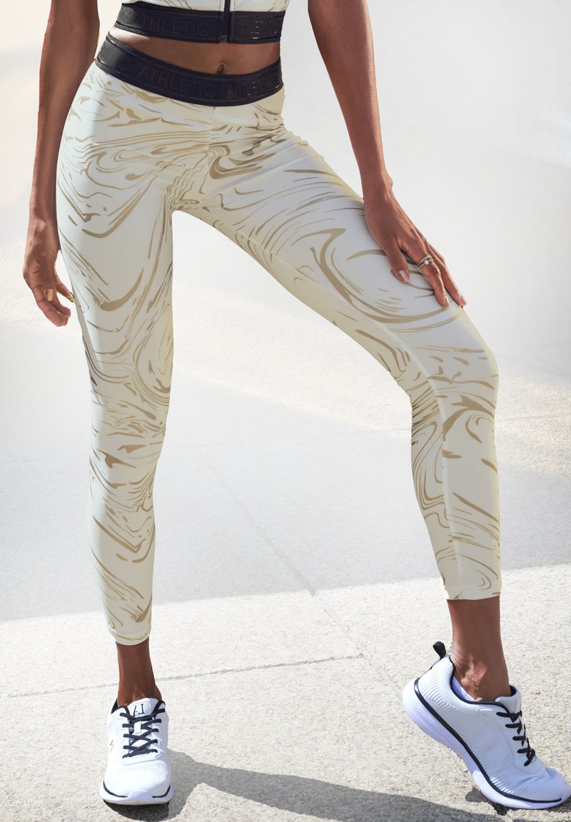 Damen Sportleggings in Creme mit goldenem Marmor-Muster, hohem Bund und feuchtigkeitsableitendem Material. Kombiniert mit weißen und schwarzen Sneakers.