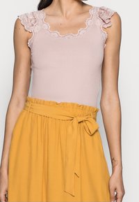 Kvinna i en ärmlös topp i rosa spets, instoppad i en senapsgul kjol med en knytbar midjeband och volangmidja.