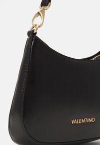 Black leather shoulder bag with curved design, gold hardware, and embossed logo. Features a zipper and smooth texture.