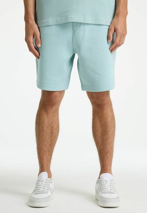 Mann trägt hellblaue Shorts und weiße Turnschuhe, steht mit entspannt an den Seiten hängenden Händen vor einem einfarbigen weißen Hintergrund.