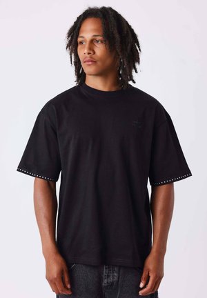 Jeune homme à la peau mate et aux dreadlocks portant un t-shirt noir ample avec de petits détails ronds le long des bords des manches.