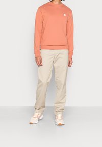 Koraalkleurige sweatshirt met een ronde hals en klein logo, gecombineerd met beige broek en lichte sneakers. Soepele stof en een relaxte pasvorm.