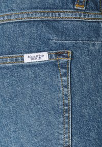 Modré džínové kalhoty s žlutým prošíváním, se zapínací kapsou a štítkem s nápisem "Marc O'Polo DENIM." Viditelné detaily strukturované tkaniny.