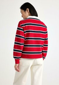 Rød stribet pullover med marineblå og hvide striber, der har en hvid krave og let puffede ærmer, båret med cremede bukser.