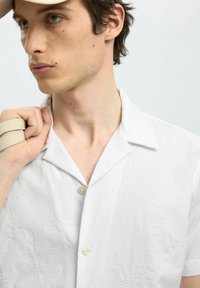 Bílé tričko s krátkým rukávem a límečkem, zdobené reliéfními květinovými vzory a jemnou texturou, zapínané na dva knoflíky vpředu.