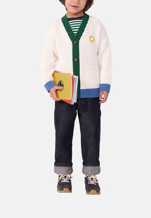 Child wearing a white cardigan with green and blue trim, dark jeans, and sneakers, holding colorful notebooks against a plain background.