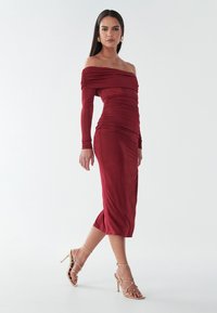 Červené midi šaty bez ramienok s dlhými rukávmi, so nariasenou textúrou a detailmi riasenia. Doplnené zlatými sandálmi na vysokom opätku.