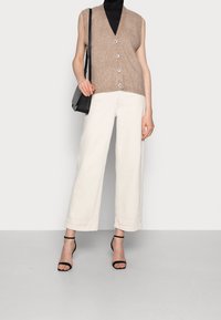 Veste sans manches en maille marron clair avec un col en V et des boutons blancs, portée avec un pantalon large blanc et des talons noirs à brides. Sac à épaule noir.