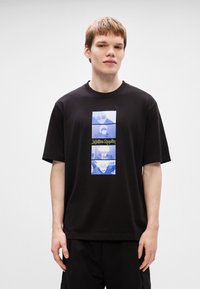 Μαύρο βαμβα κερό T-shirt με κεντρική εκτύπωση μπλε και λευκής εικόνας και στυλιζαρισμένο κείμενο, κοντά μανίκια και χαλαρή γραμμή.