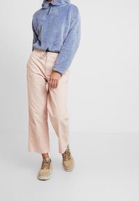 Osoba v světle fialové chlupaté mikině s kapucí, béžových širokých kalhotách a hnědých turistických botách stojící na bílém pozad�í.