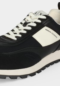 Baskets Armani Exchange noir et blanc avec lacets blancs, empeigne texturée, marquage sur le panneau latéral et la languette, et semelle extérieure crantée.