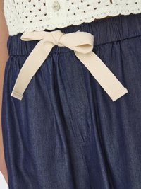 Denim broek met een donkerblauwe glans heeft een elastische tailleband en een crèmekleurige stoffen strik aan de voorkant, wat detail aan het ontwerp toevoegt.