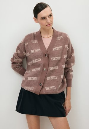Vrouw met donker, naar achteren gestyled haar draagt een mauve gebreide cardigan met knopen en "BIZUU"-print en een zwarte rok, staand tegen een effen achtergrond.