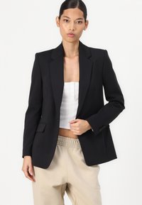 Blazer preto com um corte estruturado, lapelas recortadas e fecho de botão único. Combinado com um top branco ajustado e calças bege.