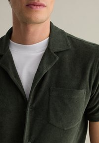 Korte groene badstof shirt met een ingesneden kraag, een borstzak en knopen. G gedragen over een wit crewneck t-shirt. Zachte textuur.