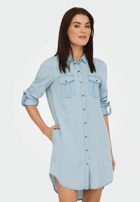 Ljusblå denimklänning med knäppning fram, bröstfickor, rullade ärmar och en svagt böjd nederkant.