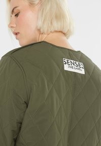 Blonde Frau, die eine olivgrüne, gesteppte Jacke mit einem "SENSES THE LABEL"-Aufnäher auf dem oberen Rücken trägt, seitlich vor einem weißen Hintergrund steht.