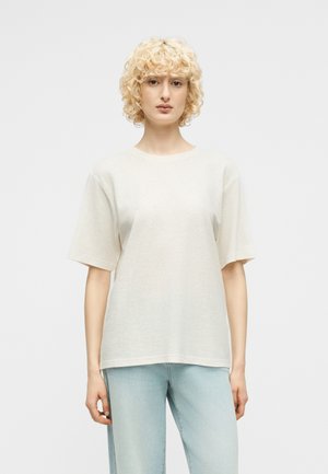 Blond osoba s kučeravými vlasmi, oblečená v jednofarebnom krémovobielom tričku s krátkym rukávom a svetlomodrých džínsoch, stojaca pred jednoduchým pozadím.