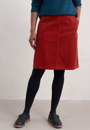 Osoba ubrana w morski sweter, rudą sztruksową spódnicę z kieszeniami, czarne rajstopy i czarne mokasyny na sprzączkę, stojąca na jednolitym tle.