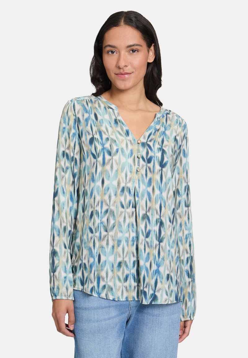 Femme portant un blouse à manches longues bleue, blanche et beige avec trois boutons, et un jean bleu clair, se tenant devant un fond blanc.