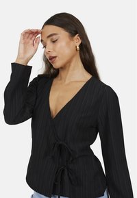 Blusa avvolgente nera con maniche lunghe, texture a righe verticali, scollo a V e dettaglio a fiocco in vita. Una leggera lucentezza esalta l'aspetto del tessuto.