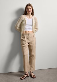 Weißes, kurzes Tanktop, leichte Strickjacke mit Kordelzügen, beige Leinenhose und schwarze Riemchensandalen auf einem grauen Boden.